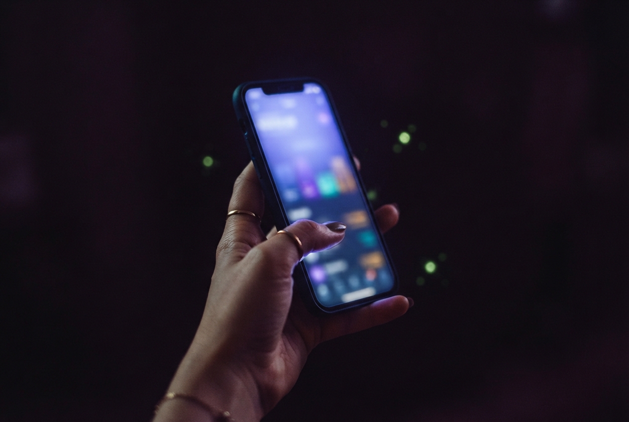 Hand with rings holding a glowing, blurred smartphone against a dark bokeh background.